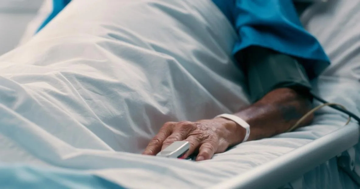 An elderly male patient lies in a hospital bed with a pulse oximeter on his finger and a blood pressure cuff on his arm.