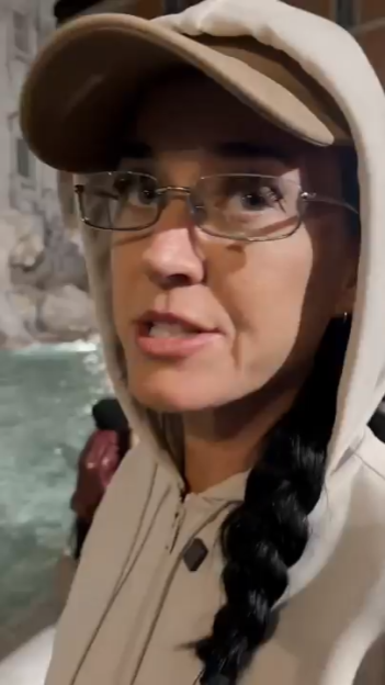 Katy Perry in a baseball cap, glasses, and a hoodie, with the Trevi Fountain in the background.