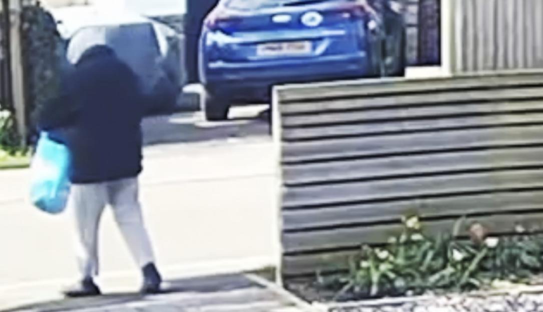 A person in a dark coat and light pants walks away from the camera, carrying a light blue bag.