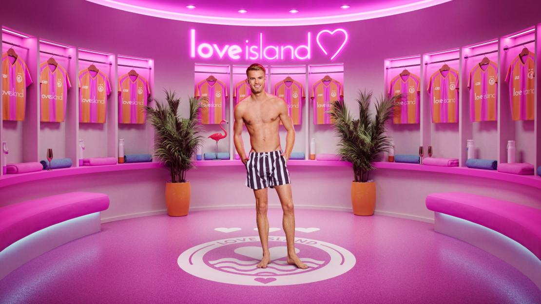 A man in swim shorts stands in a pink Love Island themed locker room, with Love Island shirts hanging in lockers.