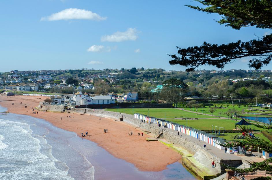 Goodrington Sands beach near Paignton, Devon, with colorful beach huts and people walking on the sand and nearby path.