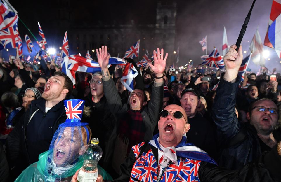A crowd of pro-Brexit supporters celebrating, many holding Union Jack flags and wearing Union Jack clothing, with some cheering with arms raised.