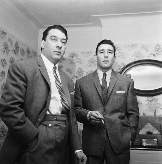 The Kray Twins, Ronald and Reginald, in suits standing at home.