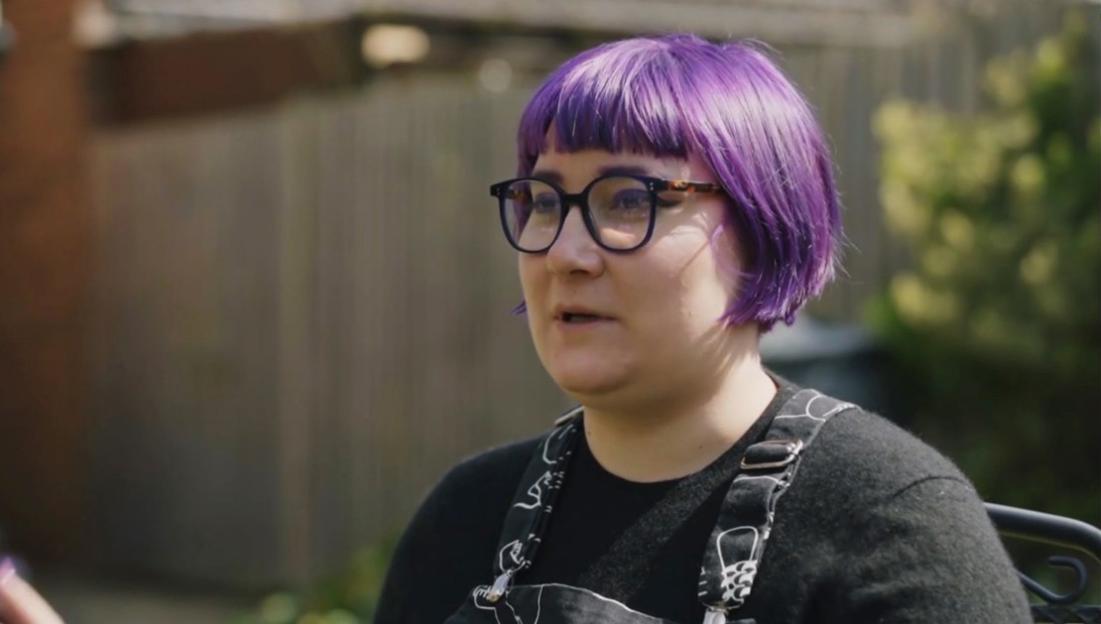 A woman with short purple hair and glasses speaks, wearing a black shirt and black overalls with white designs.