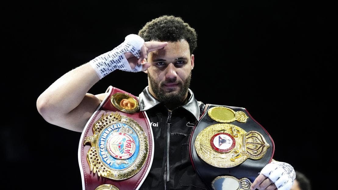 Moses Itauma, wearing two championship belts, salutes with his bandaged hand.