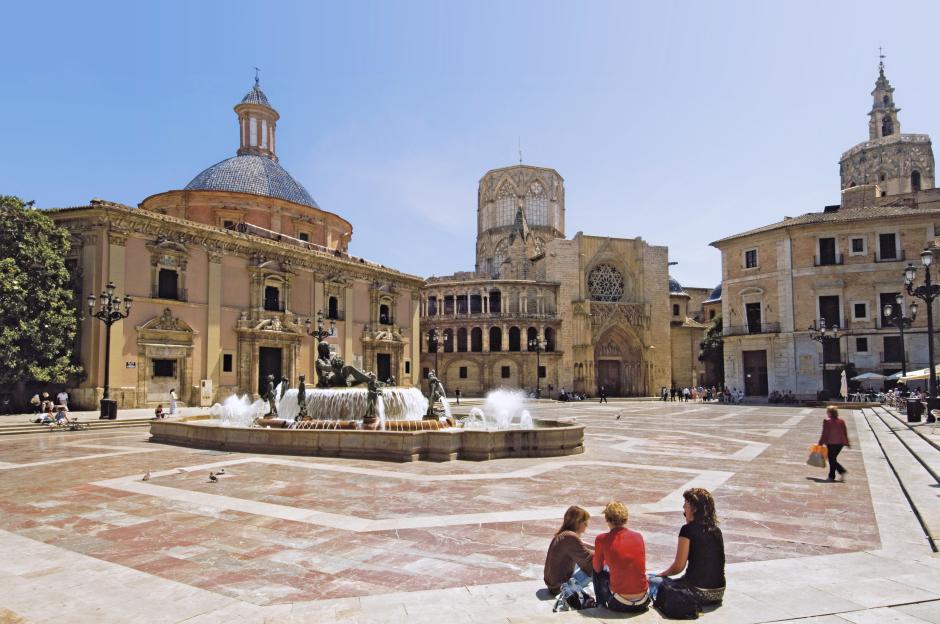Plaça de la Mare de Déu square in Valencia, Spain, featuring the Fountain of Turia, surrounding buildings, and three people sitting in the foreground.