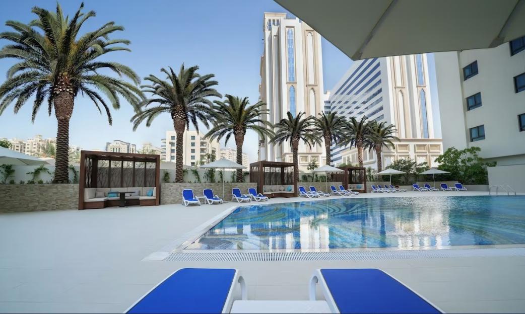 An outdoor swimming pool with lounge chairs, cabanas, palm trees, and tall buildings in the background.
