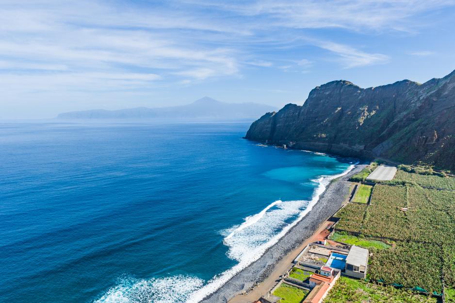 Aerial view of the beach at La Gomera island, Canary Islands, Spain, showing the blue ocean, a rocky shore, buildings, and green fields near steep hills.
