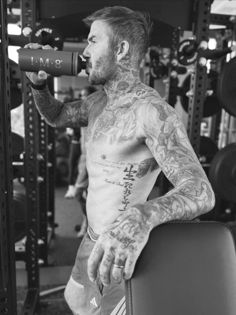 David Beckham, shirtless and heavily tattooed, drinks from an I·M·8 water bottle in his home gym.