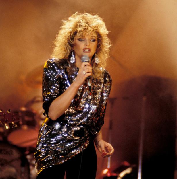 Photo of Bonnie Tyler singing into a microphone, wearing a sequined top and large earrings.