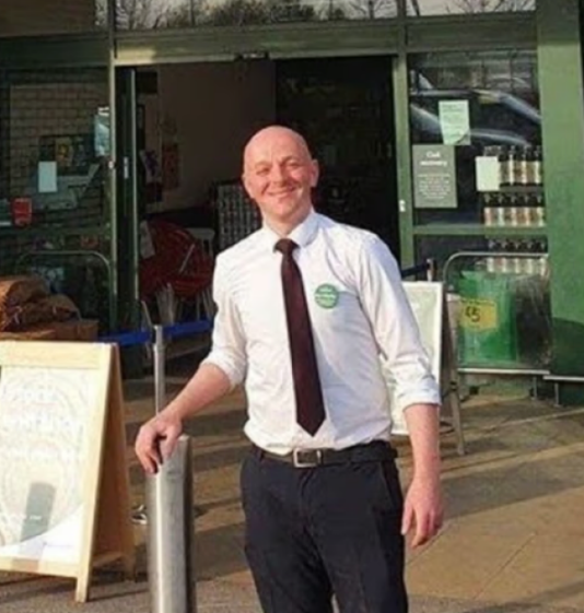 A smiling man in a white shirt and dark tie stands outside a store.