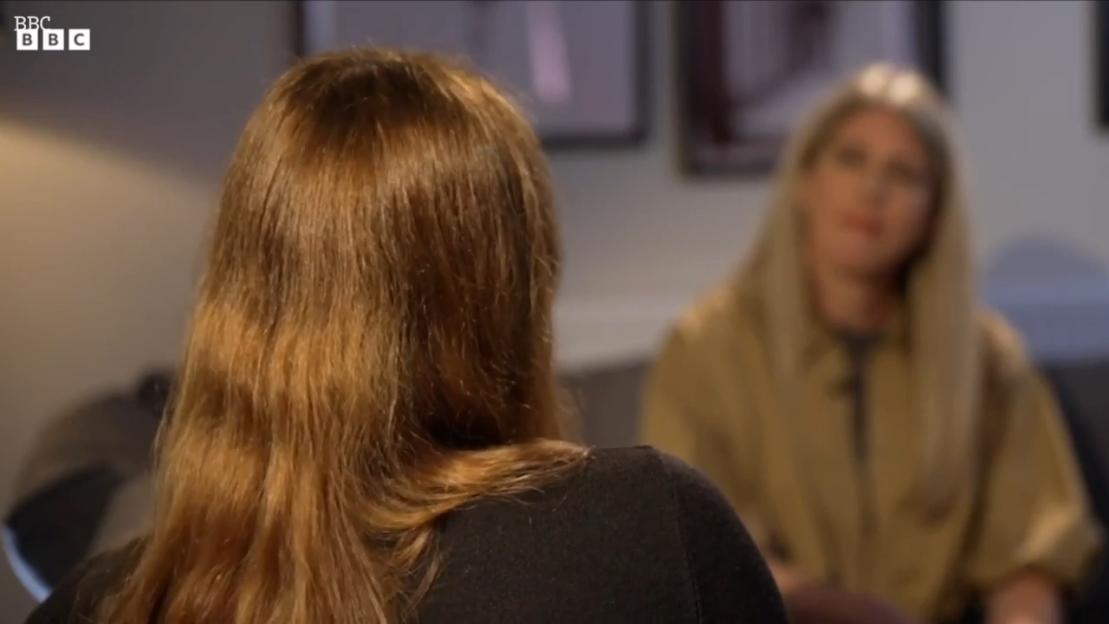 Two women, one with long auburn hair facing away from the camera, and another with long blonde hair facing the camera, are in an interview.