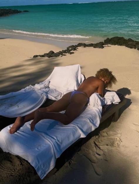 A woman in a blue bikini lies on a lounge chair on a sandy beach next to the ocean.