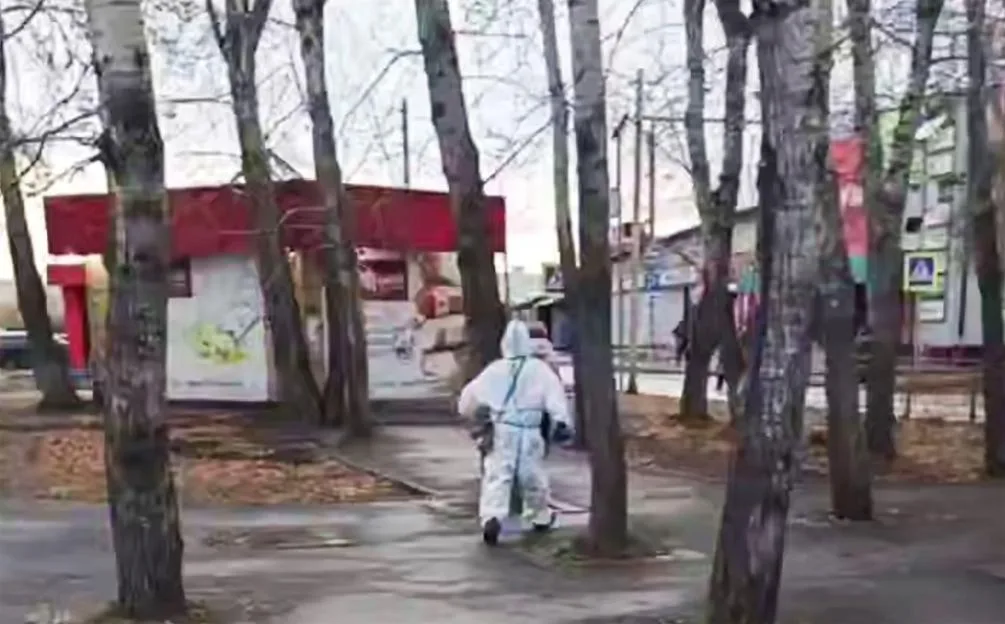 A worker in full protective gear walks past trees on a paved path in Shelekhov, Russia.