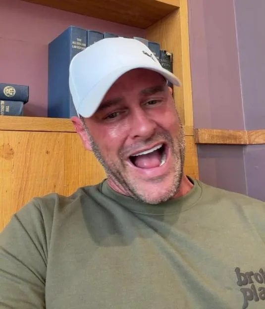 A man wearing a white baseball cap and an olive green t-shirt smiles widely with his mouth open, in front of a wooden bookshelf lined with law books.