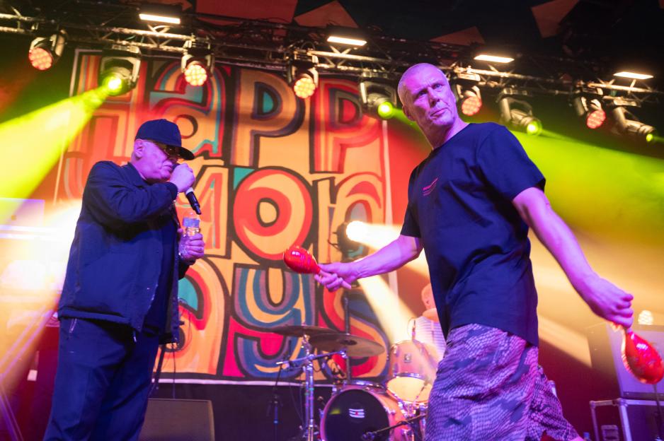 Shaun Ryder singing into a microphone and Bez shaking maracas onstage during a Happy Mondays concert.