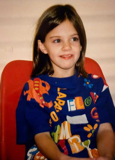 A young Chloe May sits in a red chair, looking right, wearing a blue shirt with colorful letters and designs.