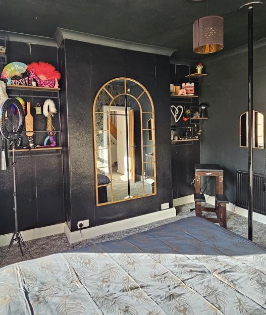 A room with black walls, a blue bedspread, a golden arched mirror, and shelves displaying various fetish items and a paddle.