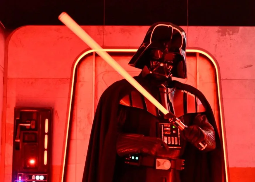 A Darth Vader costumed character holding a lit lightsaber in front of a red-lit background.