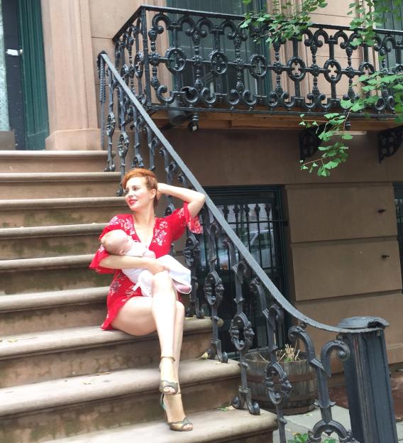 A red-haired woman in a red dress sits on stone steps, holding a baby, next to a black iron railing.