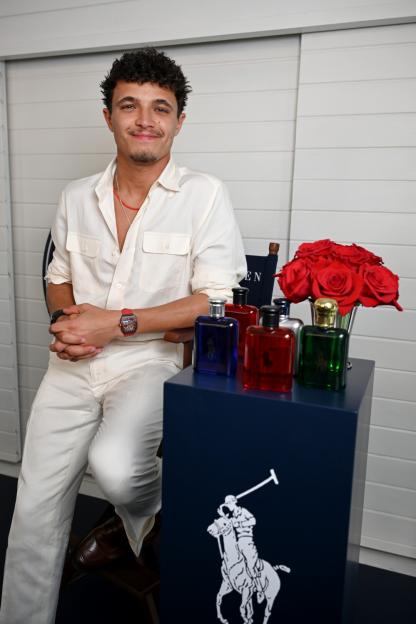 Lando Norris, wearing a cream outfit, smiles at the camera while sitting next to a blue stand displaying Ralph Lauren fragrances and a bouquet of red roses.
