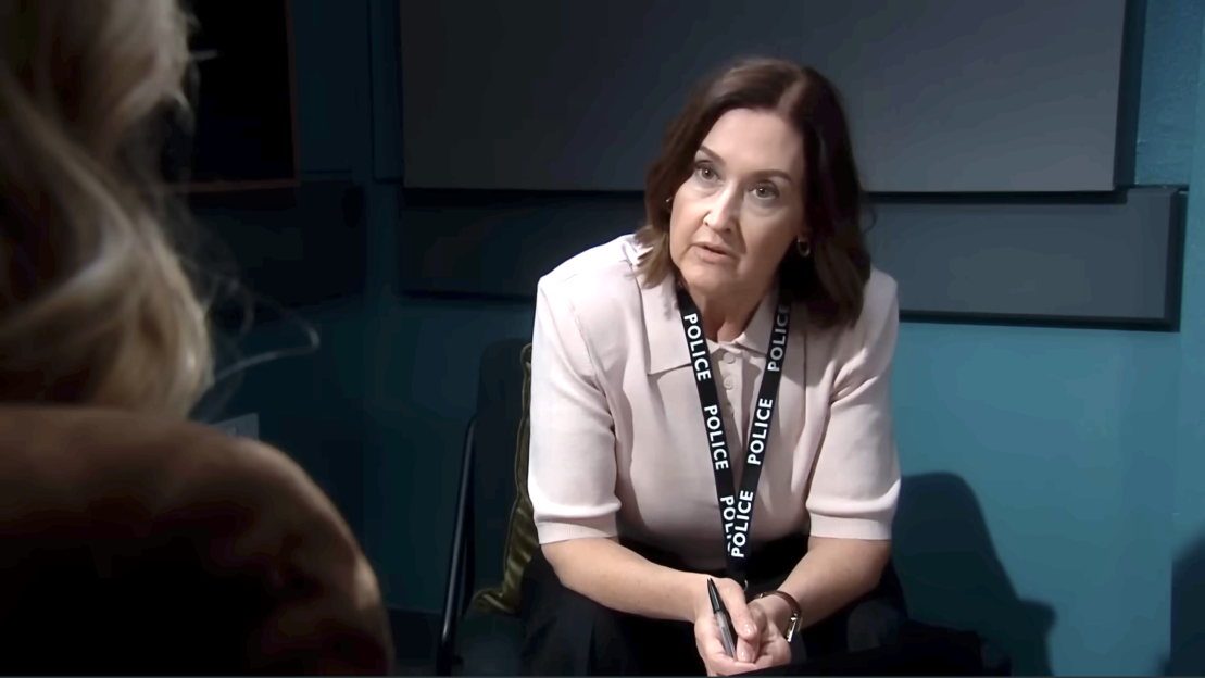 A female police officer with a serious expression, wearing a cream polo shirt and a "POLICE" lanyard, sits across from an unseen person.