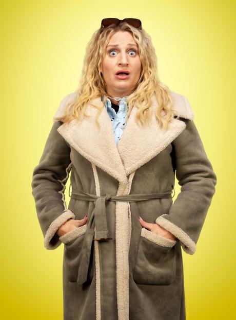 Nic (Daisy May Cooper) looks shocked and worried, wearing a brown suede coat with cream shearling lining.