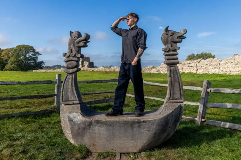 A man stands inside a wooden Viking longboat sculpture in a grassy field, saluting with one hand raised to his forehead.