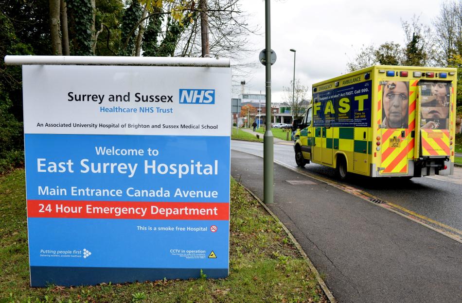A large blue and white sign for East Surrey Hospital, with a yellow ambulance driving away on a road beside it.