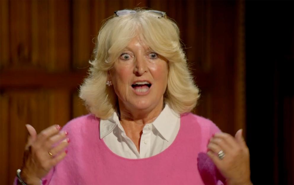 A woman in a pink sweater and white shirt looks shocked with her hands gesturing outwards.