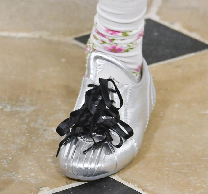 A silver sneaker with black ribbon laces and ruffled toe, worn with floral socks by Simone Rocha.