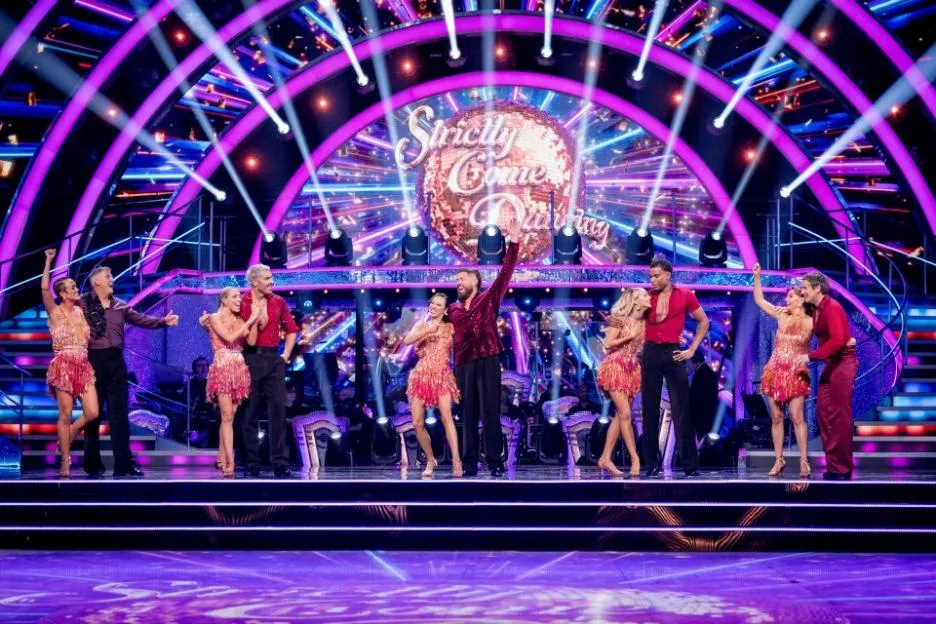 Six couples from Strictly Come Dancing, both celebrities and professional dancers, standing on stage under purple lights.
