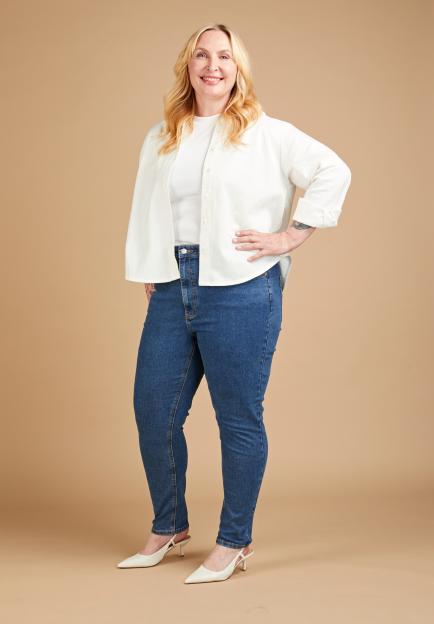 Annikki Sommerville wearing blue skinny jeans, a white ribbed top, and an unbuttoned white shirt, posing on a beige background.