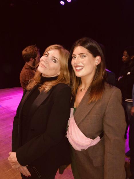 Two women, one with blonde hair and a black blazer, the other with dark hair, a brown blazer, and a pink fanny pack, stand smiling.
