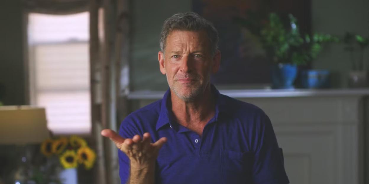 A man with short grey hair and a blue polo shirt gestures with an open hand.