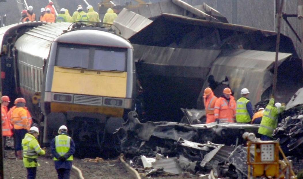 Workers in reflective gear inspect a yellow and grey train that has derailed and crashed into a building.