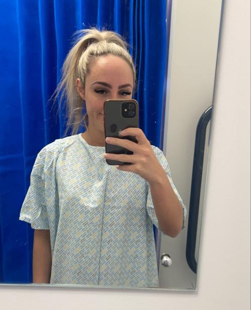 A woman wearing a hospital gown takes a mirror selfie with her phone.