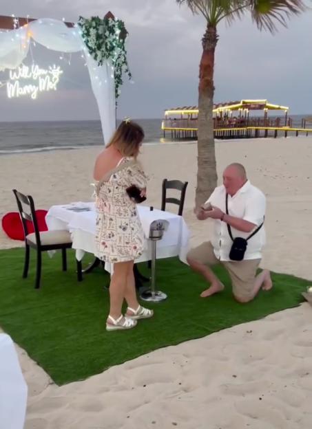 Becki Jones watches as her boyfriend Chris kneels to propose on a beach.