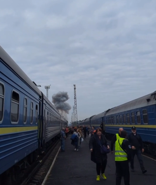 People waiting on a train platform with smoke rising in the background.