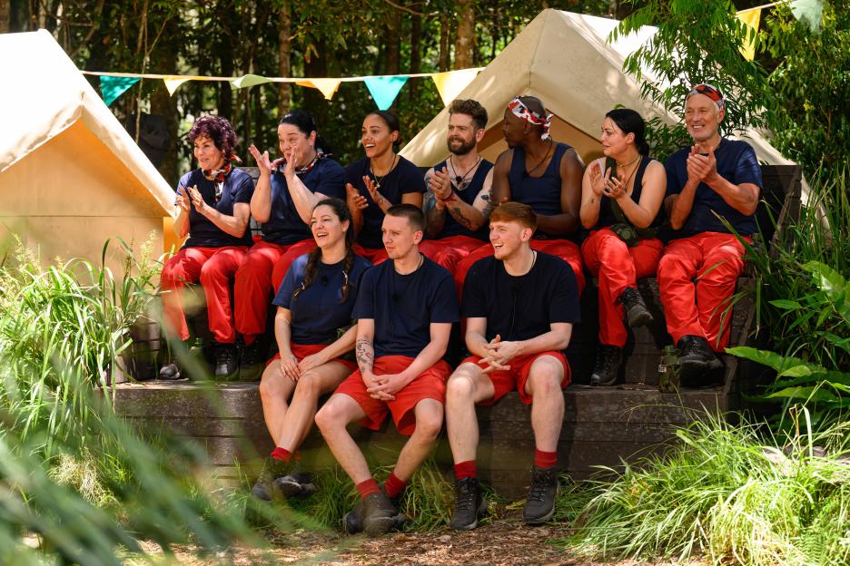 Cast members of 'I'm a Celebrity... Get Me Out of Here!' sitting on a wooden bench, clapping and smiling.