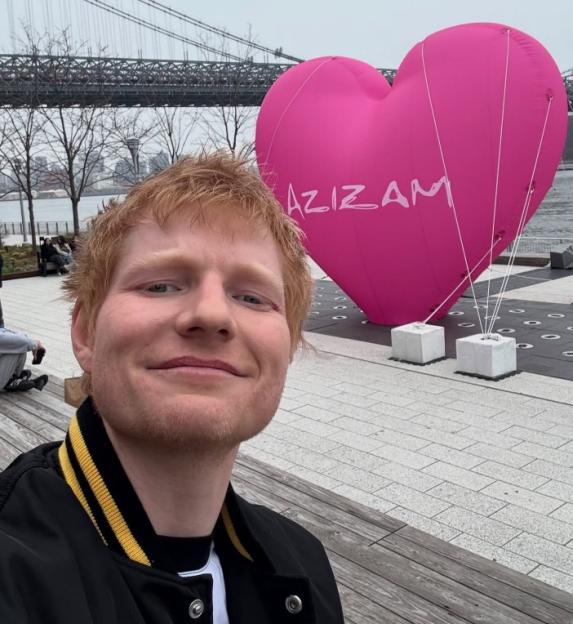 Ed Sheeran taking a selfie in front of a large pink heart balloon that says "AZIZAM".