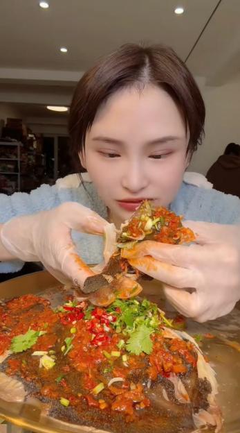 A young woman in a fuzzy blue top and clear gloves eats a spicy fish.