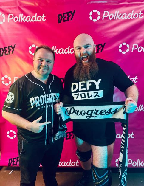 Two men smiling in front of a pink Polkadot and DEFY branded background.