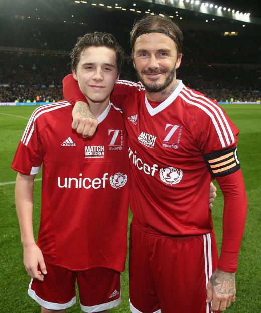 David Beckham and son Brooklyn Beckham pose in red jerseys on a soccer field after a UNICEF charity match.