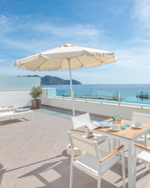 An outdoor patio at Hotel Cala Bona in Mallorca with a table, chairs, and an umbrella overlooking the ocean.