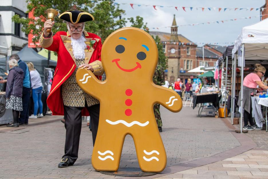A man in town crier attire holding a bell next to a large gingerbread man cutout at the Ormskirk Gingerbread Festival.