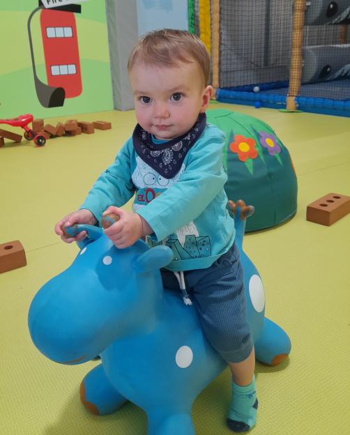 A baby boy riding a blue bouncy toy, looking at the camera.