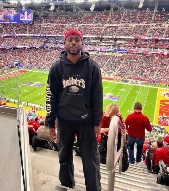 A man wearing a Las Vegas Raiders hoodie in a stadium overlooking a football field.