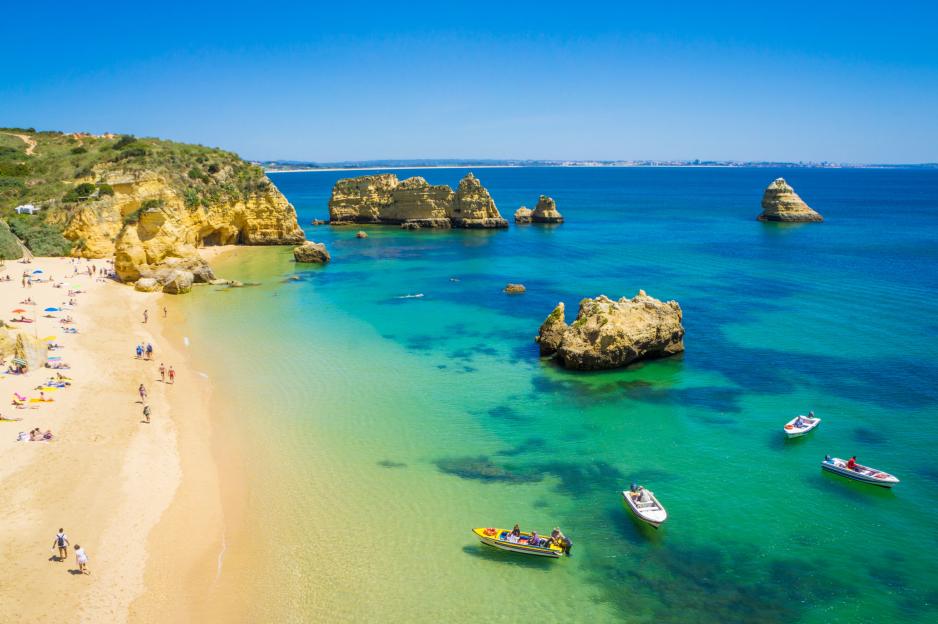 Aerial view of Praia da Dona Ana beach with sunbathers, rock formations, and boats in the clear blue-green water.