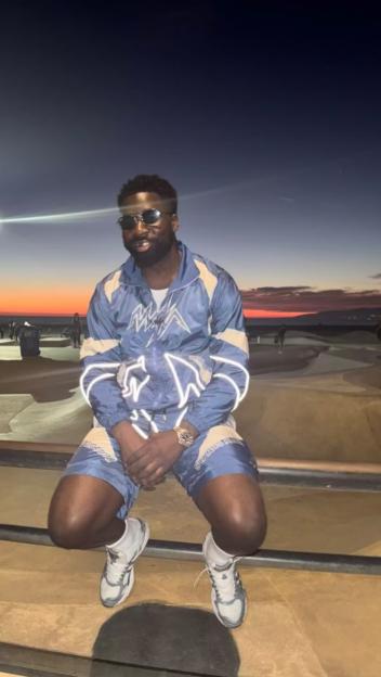 A man in a blue and white tracksuit with reflective stripes sits on a metal rail at a skate park at sunset.
